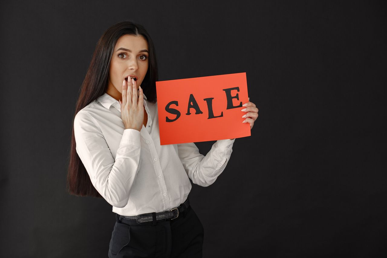 A woman in a white shirt holds a red sale sign with a surprised expression, perfect for marketing.